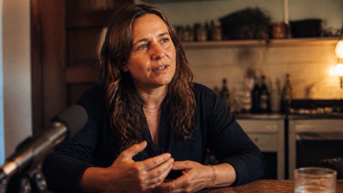 A person in their mid-forties mid-conversation at a kitchen table in warm evening light — a Legacy Decade session.