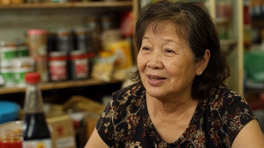 A Vietnamese-Australian woman telling her migration story in the family shop she built, warm light — the arrivals who explain why people chose here.