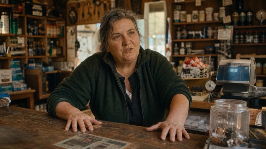 A local shopkeeper behind the counter of the shop she's run for decades, mid-story, warm afternoon light — the builders who physically made the place work.